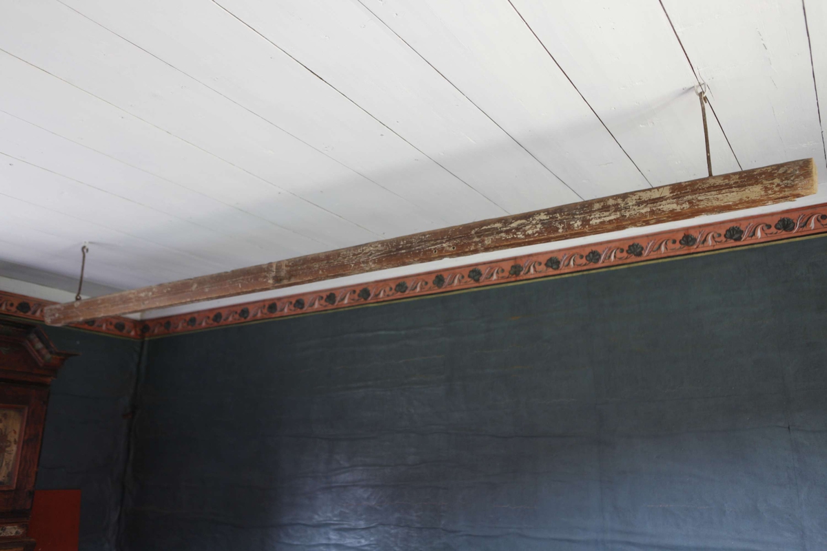 Kronstång av vitmålat trä. Rektangulärt tvärsnitt. Fäst i taket med två järnkrokar. Krokhöjd 27 cm.