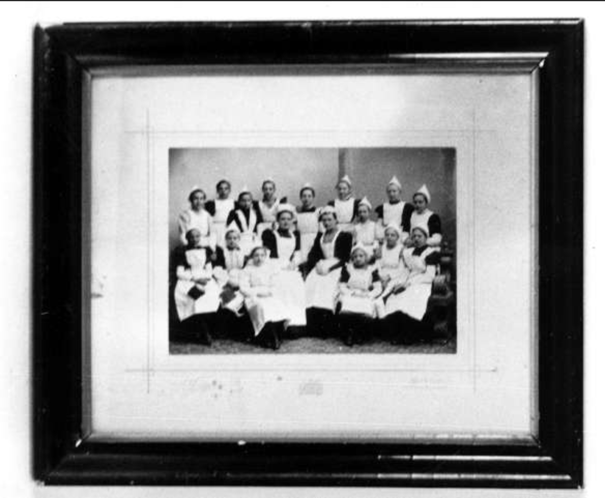 Svartvitt fotografi av skolköksklass i Uppsala - sittande flickor och två lärarinnor i vita förkläden och spetsiga mössor. 