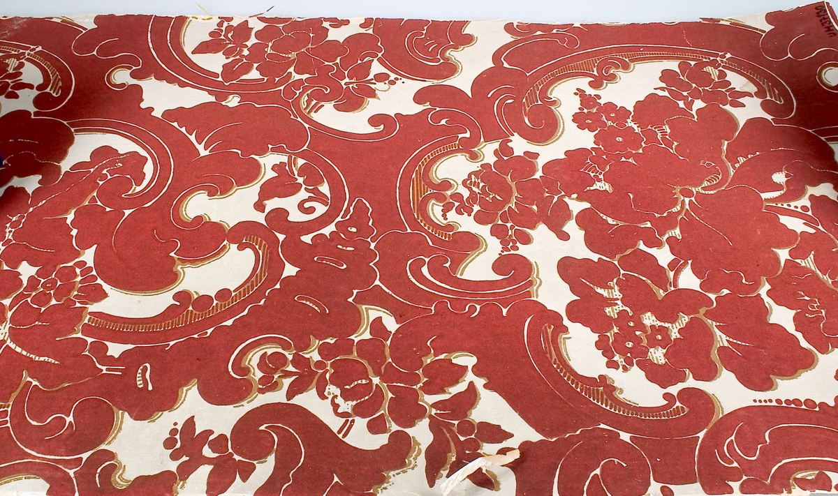 Ett tapetprov, tryck i rött och guld på vit botten. På baksidan "förstärkning" i form av rutmönster av textil - hampa eller jute.
