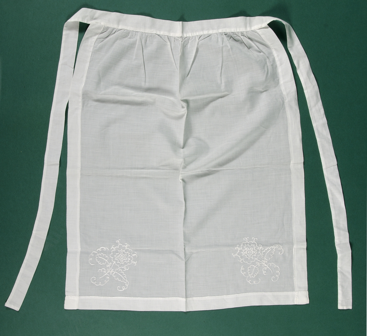 Förkläde, handsytt av tunt vitt bomullstyg vävt i tuskaft. Förklädet är fållat runt om med kedjestygn och i de nedre hörnen är blommor broderade med kedjestygn. Kedjestygnen är sydda med vitt silkesgarn.