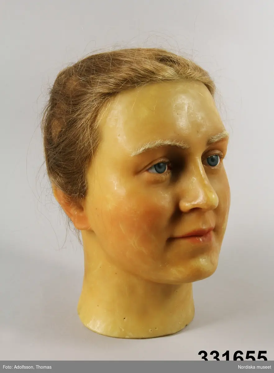 Mycket realistiskt huvud av yngre kvinna skulpterat efter levande modell. Av vax, med blå ögon och äkta cendréfärgat hår uppsatt i knut, ögonfransar och ögonbryn.
Rakt avskuren hals som kan träs på en utställningsfigur.
Anm. Spricka i nacken. repor på kinden
/Berit Eldvik 2012-10-18