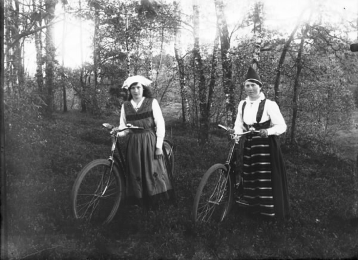 Gruppbild av två unga kvinnor ikädda folkdräkt med varsin cykel. (Se även bildnr HB194)