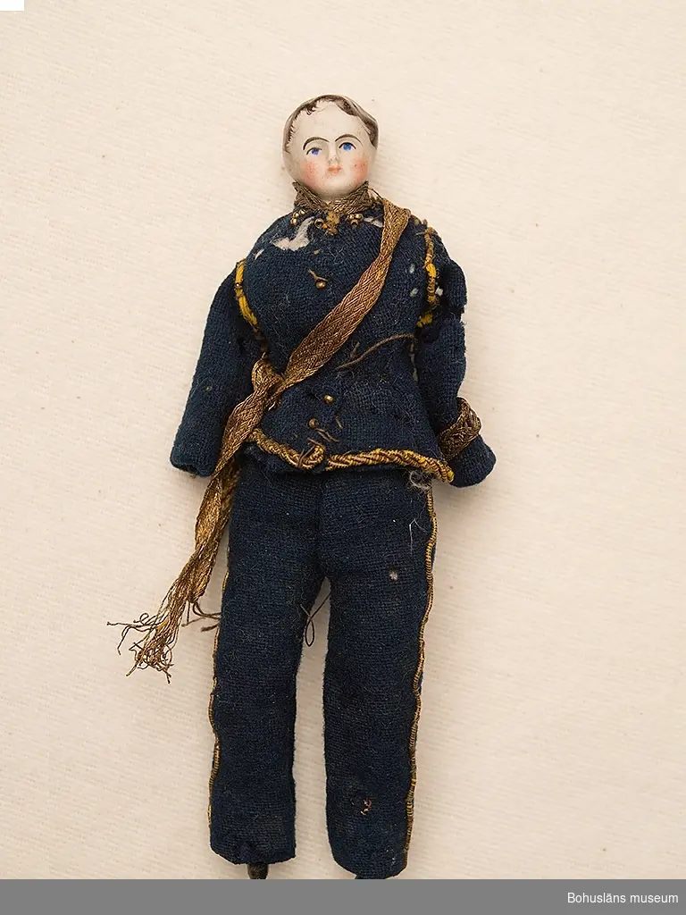 Föremålet visas i basutställningen Uddevalla genom tiderna, Bohusläns museum, Uddevalla.

Pojkdocka med tygkropp och underben och huvud i biskviporslin. Huvudet bemålat. Armar saknas. Klädd i ursprunglig uniform i blå vadmal med revärer och kanter av gul snodd. Rester av gehäng i guldtrådsliknande tyg. Jacka med skador, möjligen av mal.
Dockans kropp är hemtillverkad.