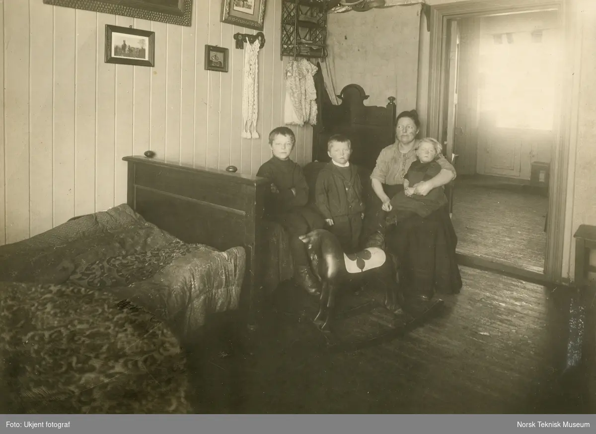Mor med tre barn fotografert i oppholdsrom i leilighet i Stavangergaten, Oslo. Bilde brukt i Barselhjemustillingen 1916