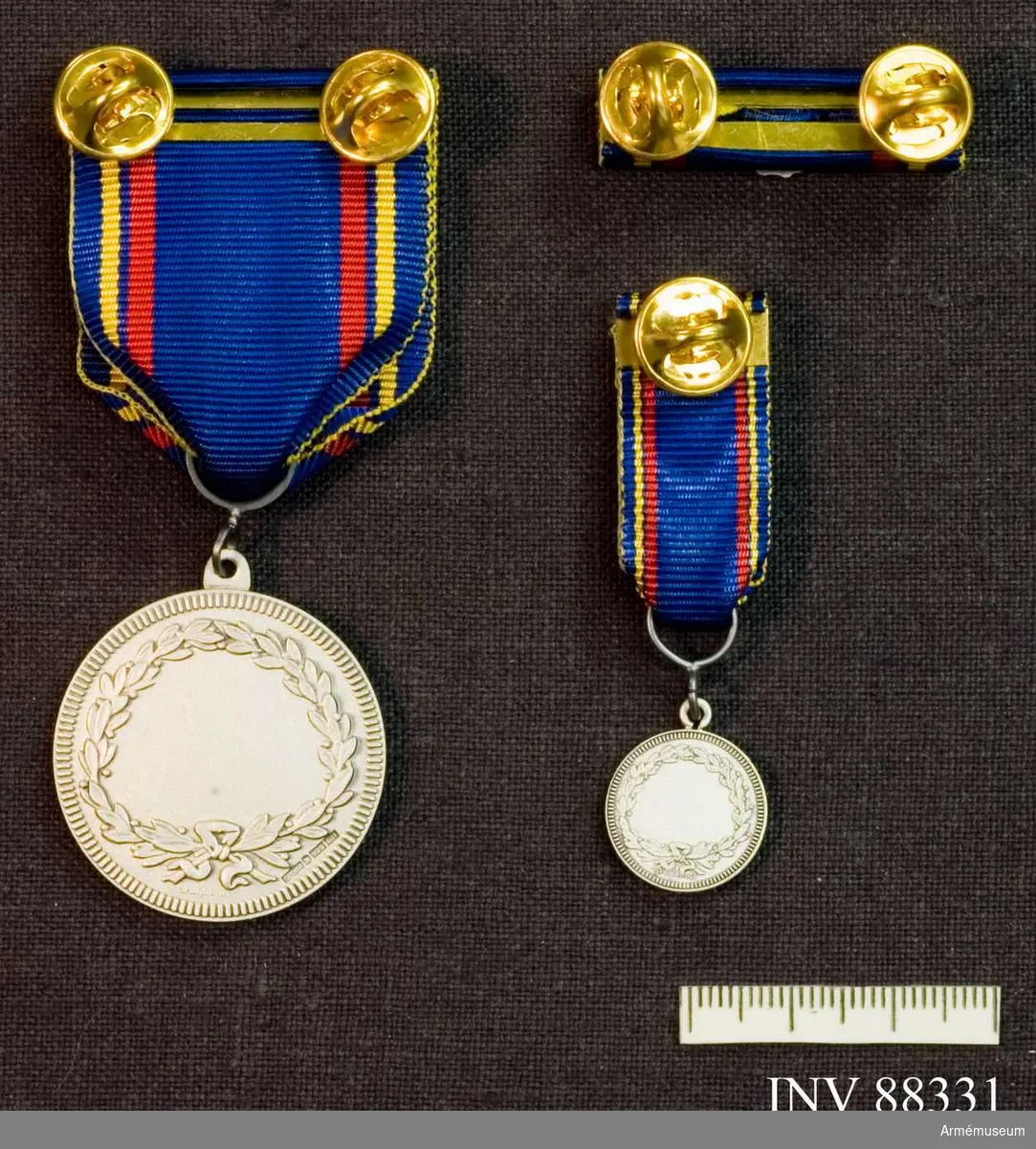 Tre samhörande föremål; en medalj, en minatyrmedalj och ett släpspänne. Bandet blått med en gul och en röd rand på vardera sidan..