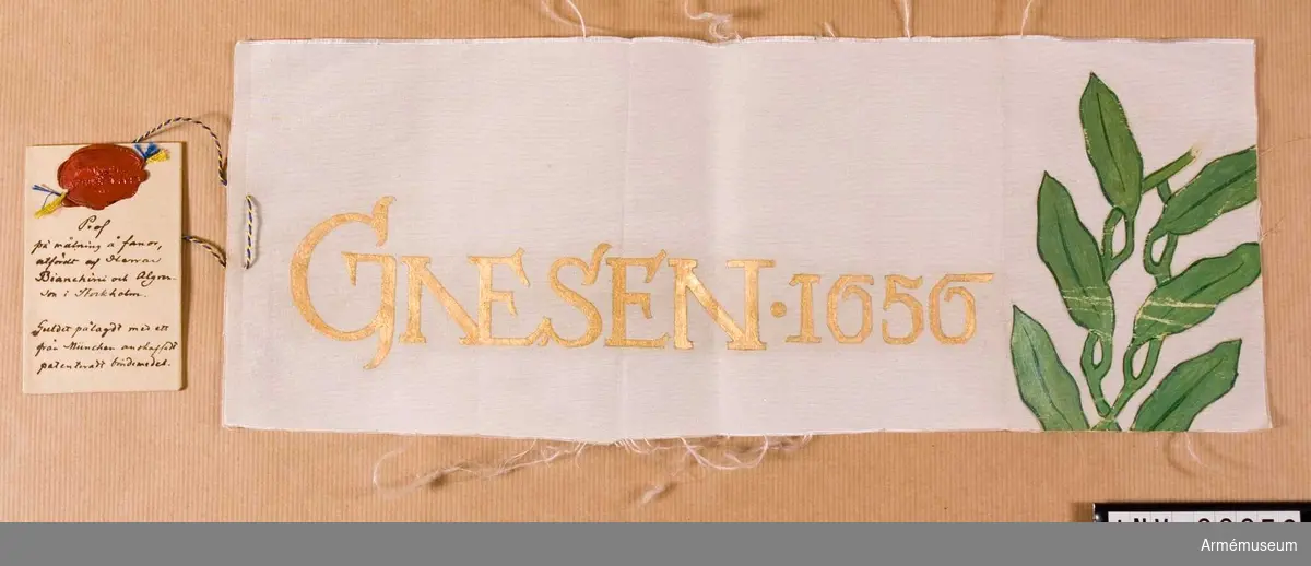 Vitt sidentyg, bemålat, med vidhängande modelletikett med text:
Prof på målning å fanor, utfördt af Herrar Bianchini och Algrenson i Stockholm. Guldet pålagt med ett från München anskaffadt patenteradt bindemedel.
