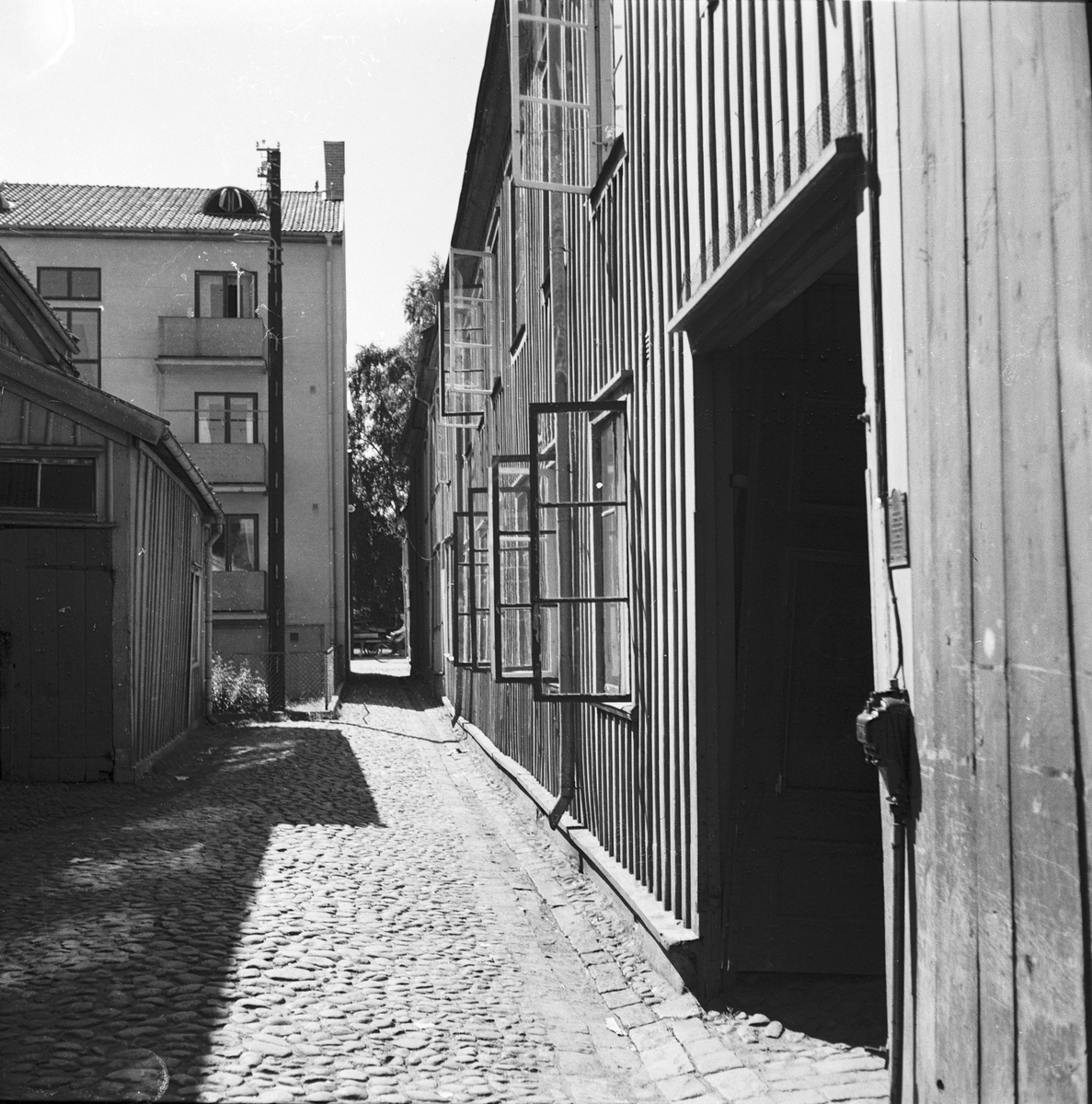 Rökerigränd i Jönköping, en kullerstensgata. Till höger ett bostadshus i trä med en port in till gården, ett antal fönster är öppna. I bakgrunden ligger ett stenhus utmed Odengatan.