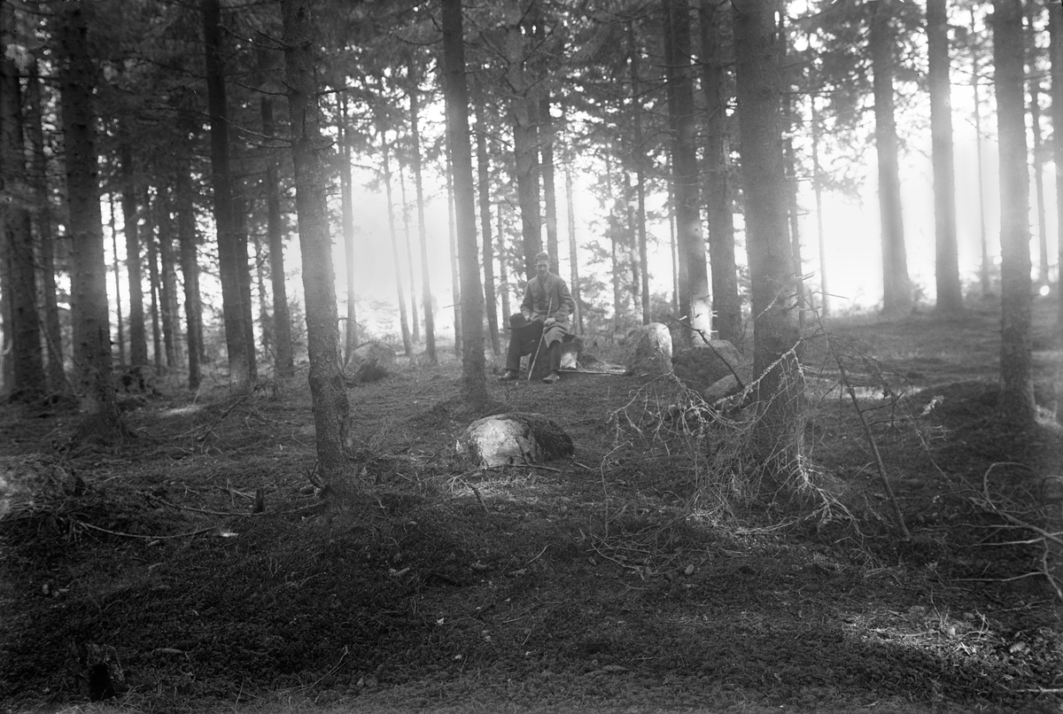 På en sten i granskogen sitter en man med en käpp i handen, sin hatt i knät och en väska bredvid sig på marken.