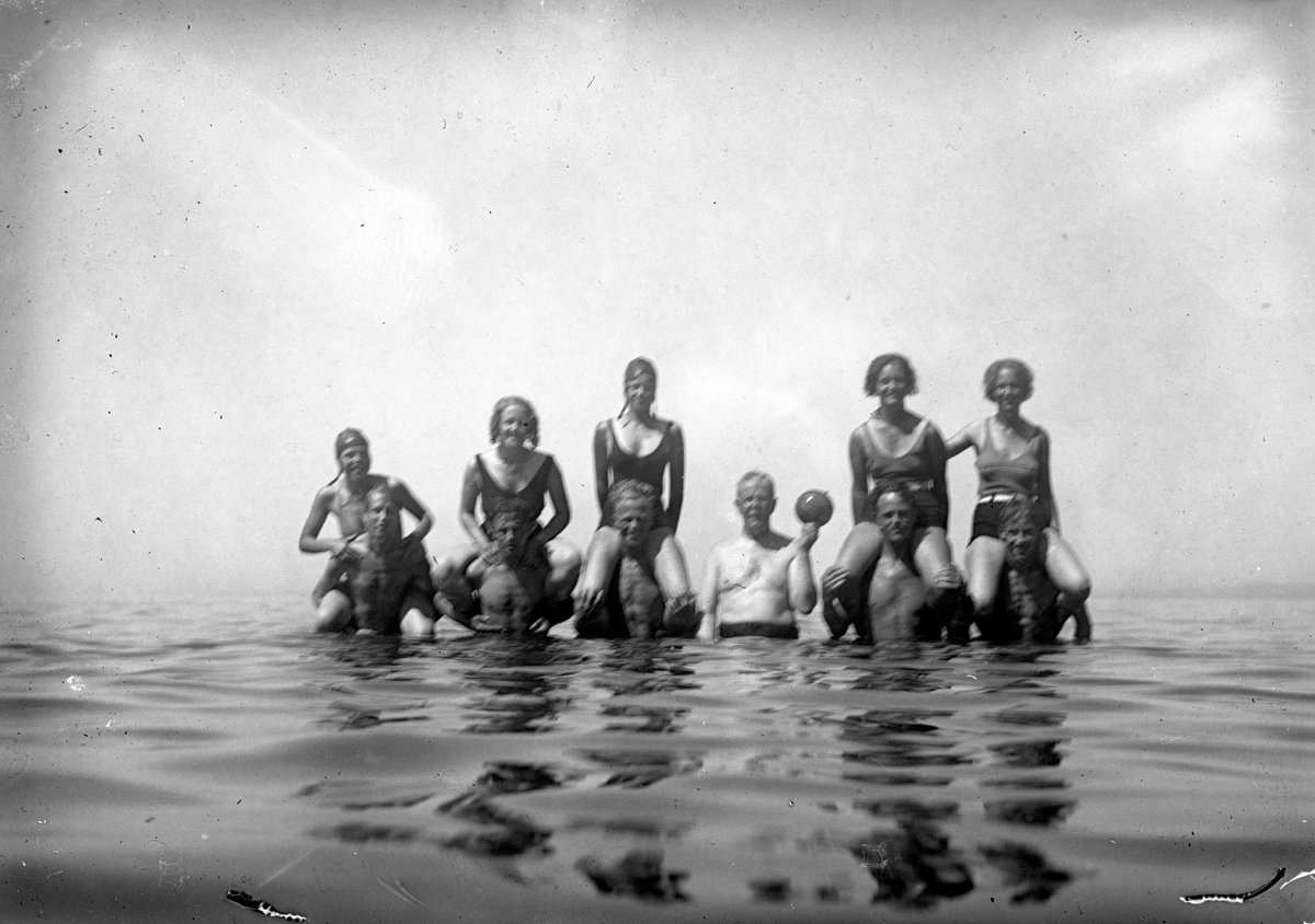 Några ungdomar badar i sjön, en man håller i en boll för vattenlek.