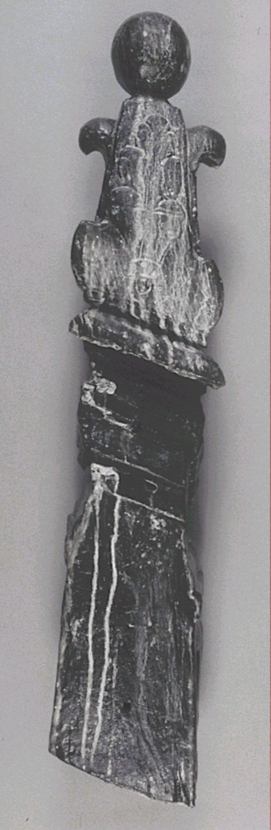 Ornament, upptill formad som en obelisk, krönt av ett stort klot.
Obelisken vilar på en profilerad och snedställd platta som avgränsar den från en hög sockel. Obelisken är skuren i ett stycke samt är rikt dekorerad med stora, långsträckta och mångflikiga blad, liknande eklöv. Bladflikarna på obeliskens kortsidor är delvis avskurna. Obelisken är dekorerad runt om vilket antyder att den varit placerad fristående.

Sockelns övre del består av ett tjockt textilstycke, format likt en scarf, som trätts genom en stor ring. Nedtill är sockeln prydd med en enkelt skuren maskaron med näbbliknande mun, omgiven av en krans med smala inskärningar. Maskaronen begränsas nedtill av en profilerad vulst. Sidorna är prydda med rullverksspiral. Sockeln har en avplanad baksida. På baksidan finns ett snedställt urtag som är försett med skåror och upphöjda åsar. På kortsidorna till obeliskens bas utgörs dekoren av en enkelt inristad rektangel.
Skulpturen är mycket välbevarad.

Text in English: Ornament in the shape of an obelisk, topped with a large sphere and placed upon a pedestal.
A plinth seperates the obelisk from the pedestal. The plinth is flattened at the back. The obelisk is carved in one piece, three-dimensionally, and is richly decorated on all four sides  with large, multi-lobed leaves similar to oak leaves. On the short sides of the obelisk''s thickened base there is a simple engraved rectangle.

The leaf lobes on the narrow sides of the obelisk are truncated. A mask, with a face with a beak-like mouth, decorates the pedestal. The mask is surrounded by a wreath with narrow incisions which terminate below in a torus. Scrollwork decorates the sides. Above the mask a festoon of carved ribbon passes through a large ring.
The sculpture is very well preserved.