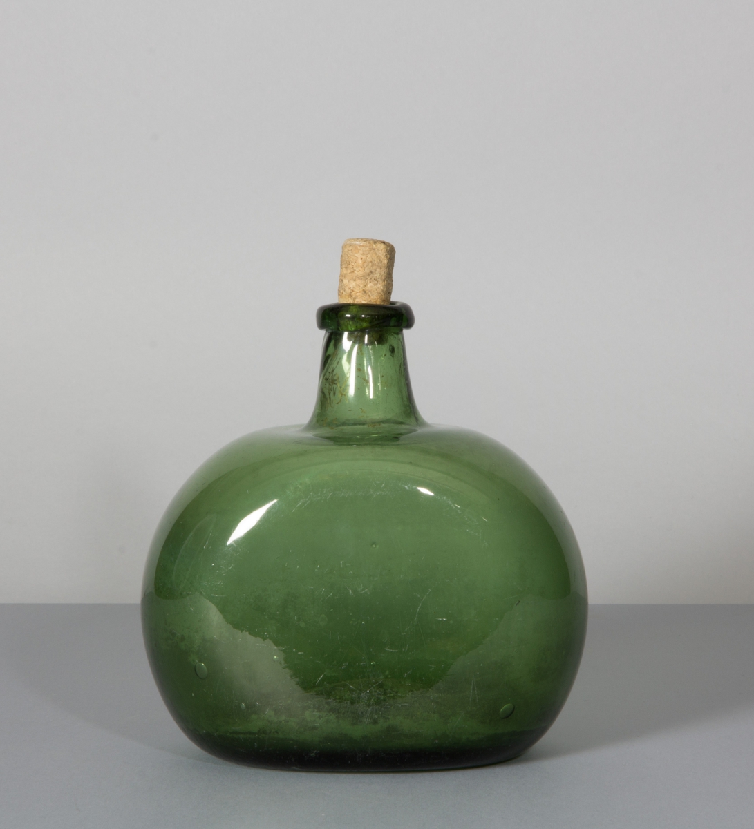 Flaska av glas, grön till färgen. Rundade sidor, hals med halsring och platt botten.