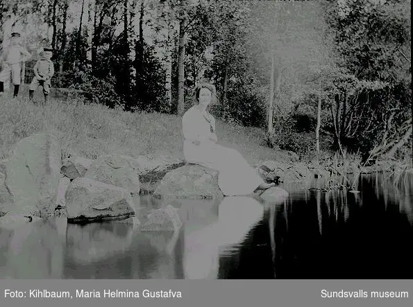 Porträtt. Kvinna sitter vid en å eller sjö, pojkar står bakom.