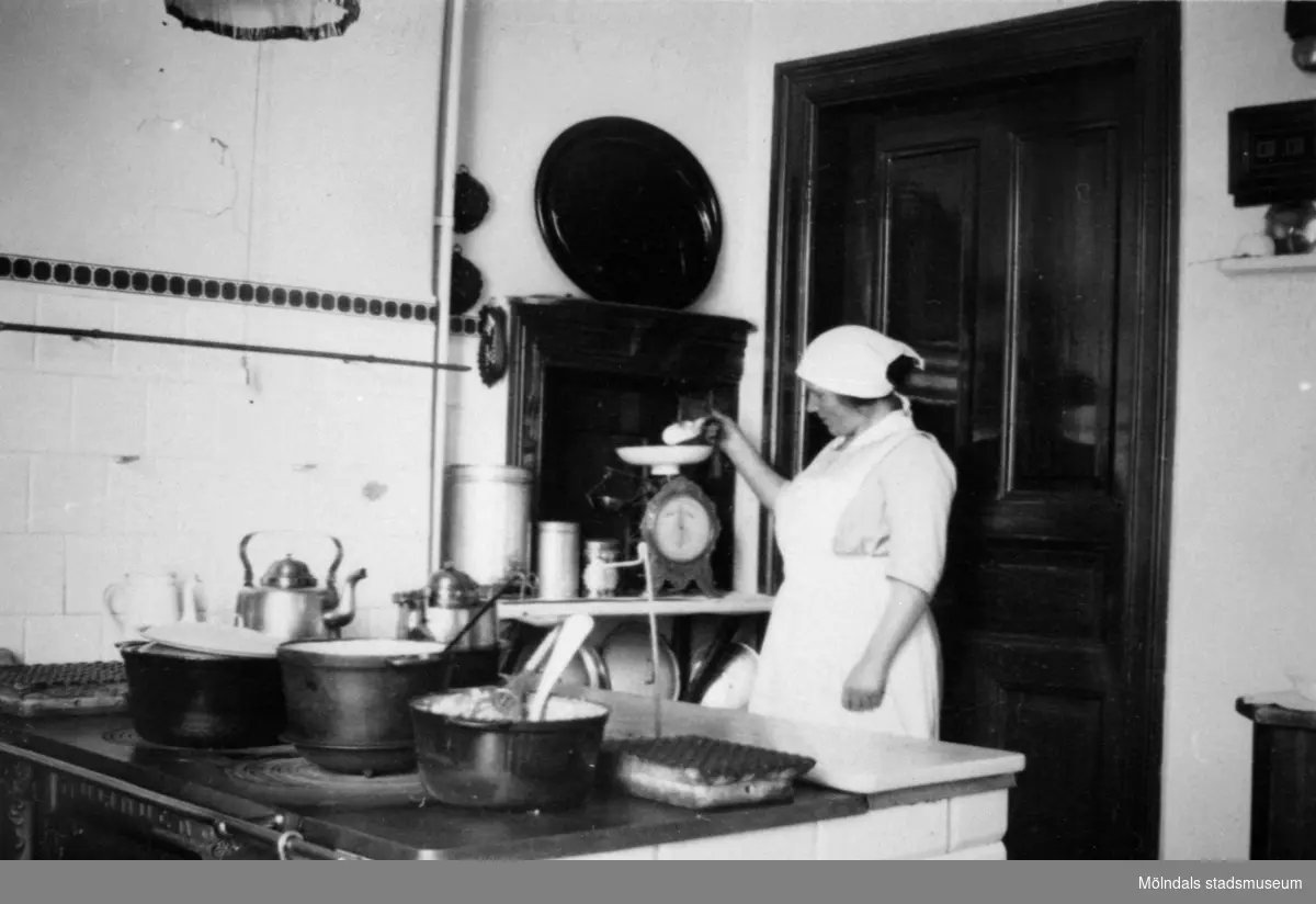 Hilma Olsson arbetar i köket och mäter upp något på en våg i köket i Stora Skolan på Stretereds skolhem okänt årtal. Hennes son (Åke Olsson, född i början av 1920-talet) var fosterson i Vommedal.