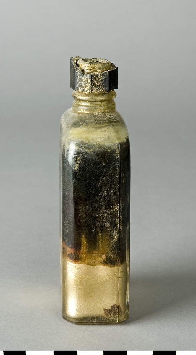 Flaska av glas med svart skruvkork av plast. Flaskan innehåller BRONSTINKTUR från Beckers. En prislapp på baksidan anger priset KR 3.75. I nedre tredjedelen av flaskan ligger färgämnet samlat.