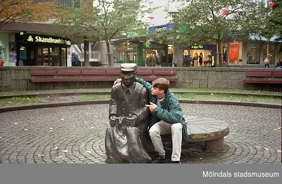 En kille sitter vid en skulptur på Alberts torg. Mölndalsbro i dag - ett skolpedagogiskt dokumentationsprojekt på Mölndals museum under oktober 1996. 1996_1227-1245 är gjorda av högstadieelever från Kvarnbyskolan 9D, grupp 6. Se även 1996_0913-0940.
