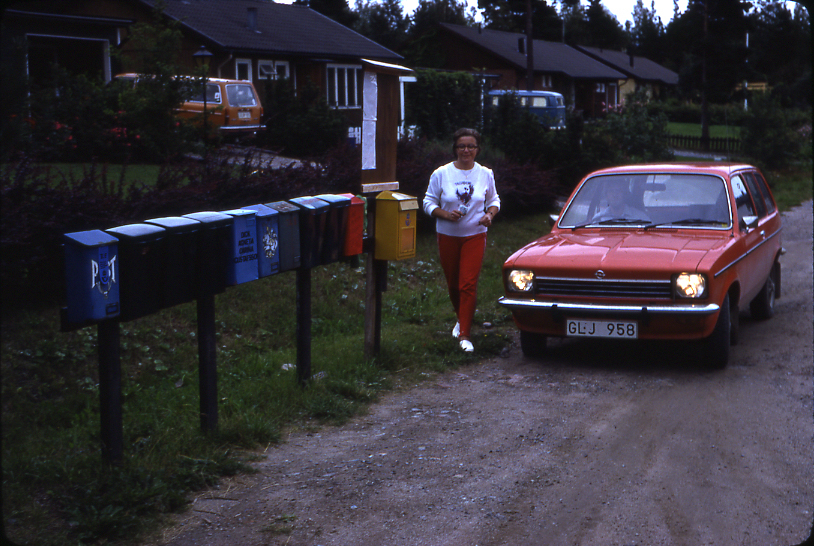 Lantbrevbärare Mikael Mattsson har kommit till Rocksta. Gunvor Wessberg går vid bilen. I förgrunden syns en samling postlådor och en gul brevlåda.