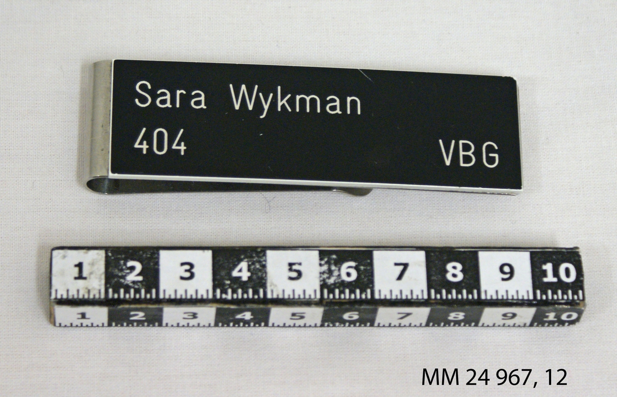 Namnbricka av stål. Rektangulär platta med clips. På plattan en svart bricka med tryckt text i vitt: "Sara Wykman 404 VBG" (Visborg). Kronstämpel på baksidan.