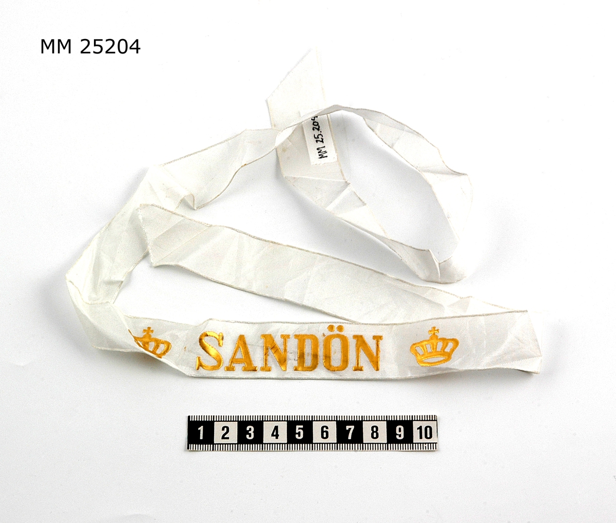 Mössband av sidenrips. Vit med text i guld: "Sandön" samt krona i guld på vardera sida om namnet.