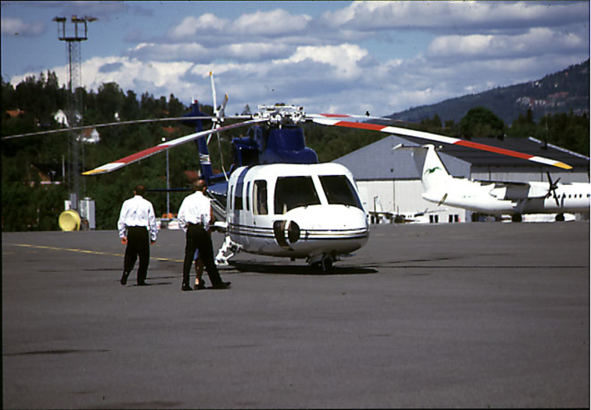 Lufthavn, 1 helikopter på bakken, Sirkorsky S-76B, N31KR fra Kjell I. Røkke. 3 personer foran helikopteret. 2 fly og flyplassbygninger i bakgrunnen.