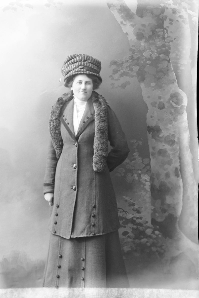Studioportrett av kvinne i helfigur,  iført hatt og pels.