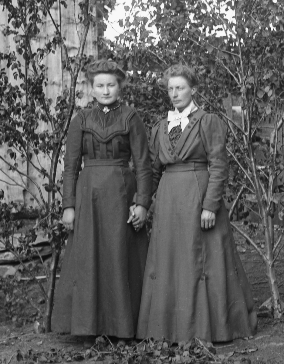 To kvinner i helfigur kledd i mørke kjoler, som holder hverandre i hånda, foran husvegg og bjørkekvister