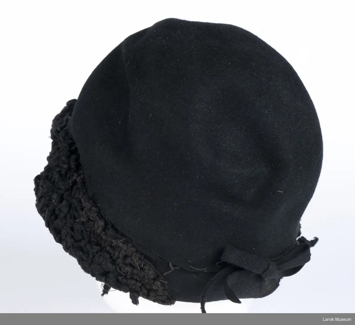 Form: sort filt, langs forsiden besetning av sort persianerimitasjon og garnityr av 5 plater perle- morsimitasjon og 2 små perler, filtsløyfe bak, sort ripsbånd på innsiden, omsydd 