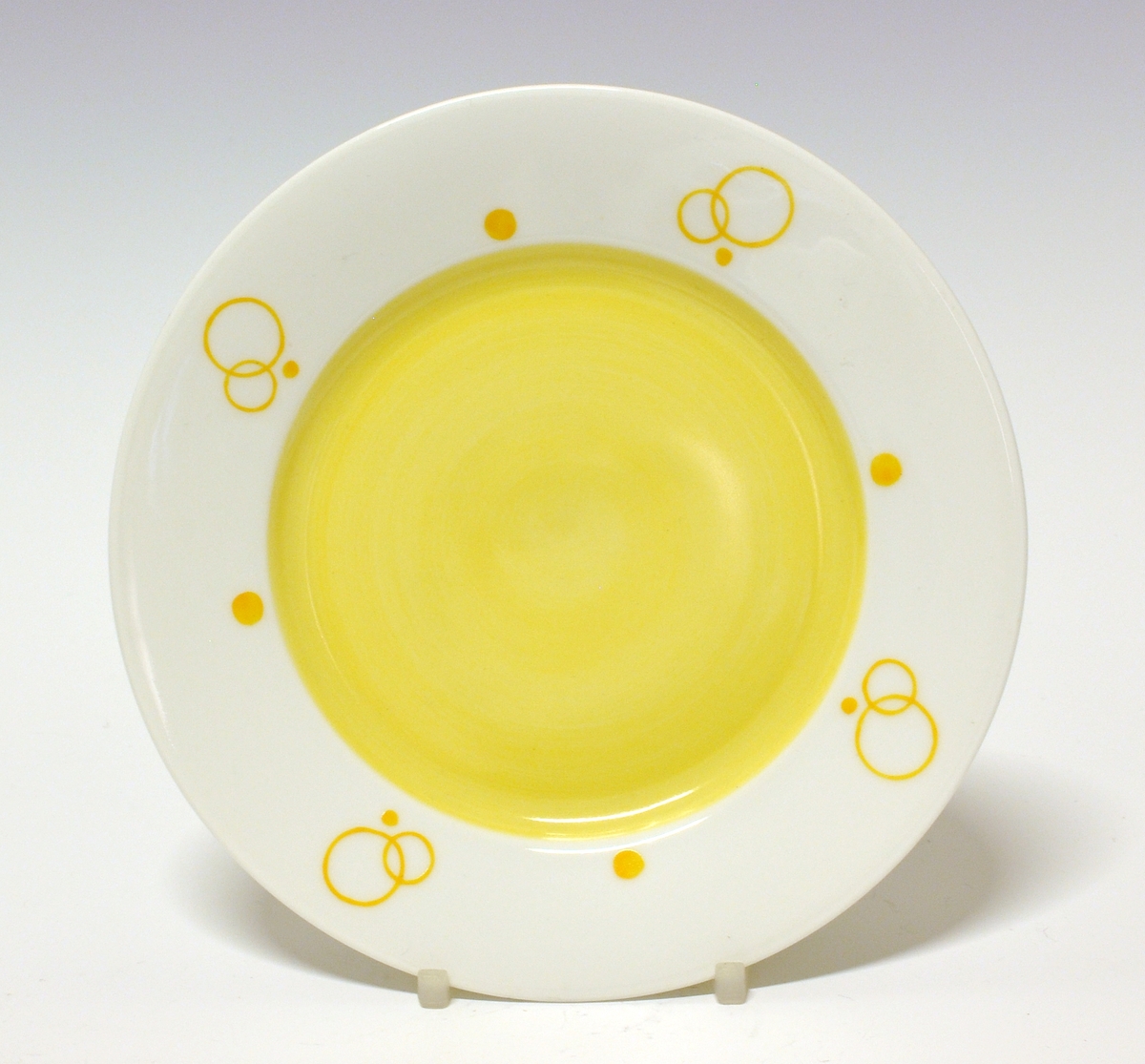 Asjett av porselen. Hvit glasur. Speilen malt gul og på fanen gule ringer og prikker.
Modell: 15.5
Dekor: 5837