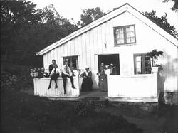 Mennsker på veranda. Barn sitter på rekkverket. Bilde er tat