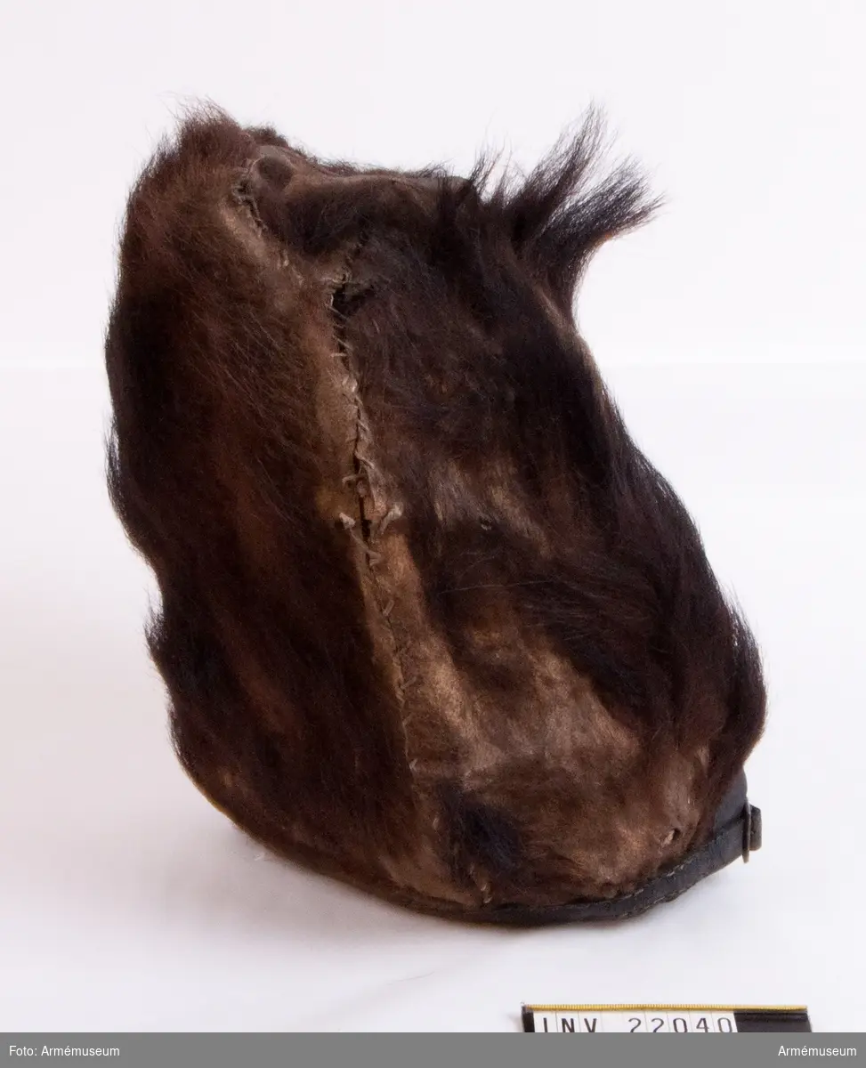 Mössan, som är helt tillverkad av björnskinn, är stoppad på en spjälram med innerhatt av filt. 
Mössan saknar brätte men har en läderrem fastsydd i nederkant med ett mässingsspänne bak.