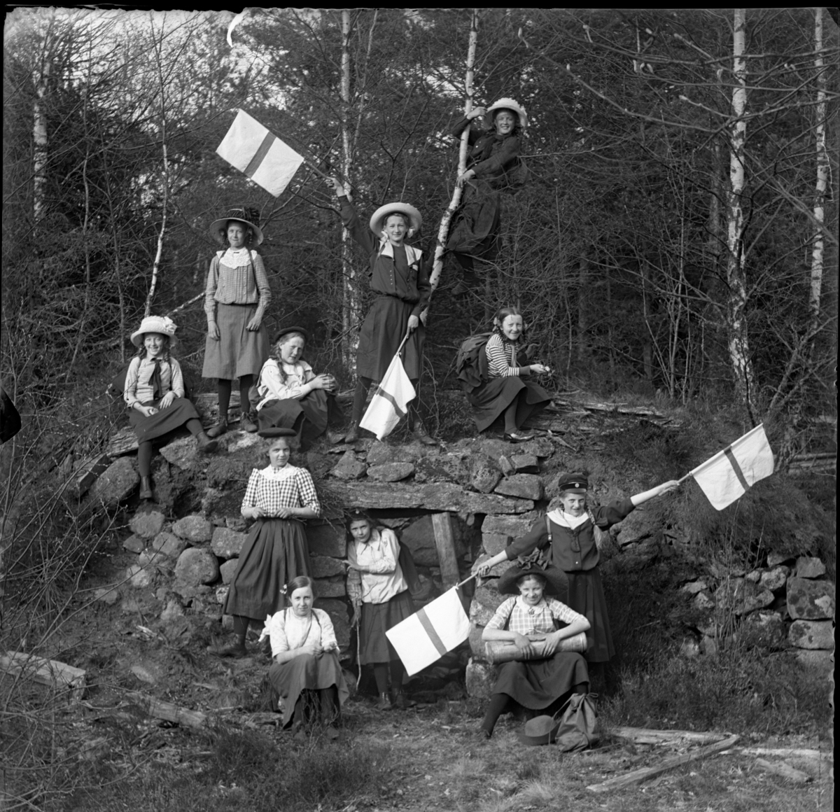'Gruppbild med 11 flickor samlade vid en jordkällare byggd av stenblock. Iklädda hatt, keps, kjol. Viftande med slags flaggor. ::  :: Ingår i serie med fotonr. 5415:1-10.'
