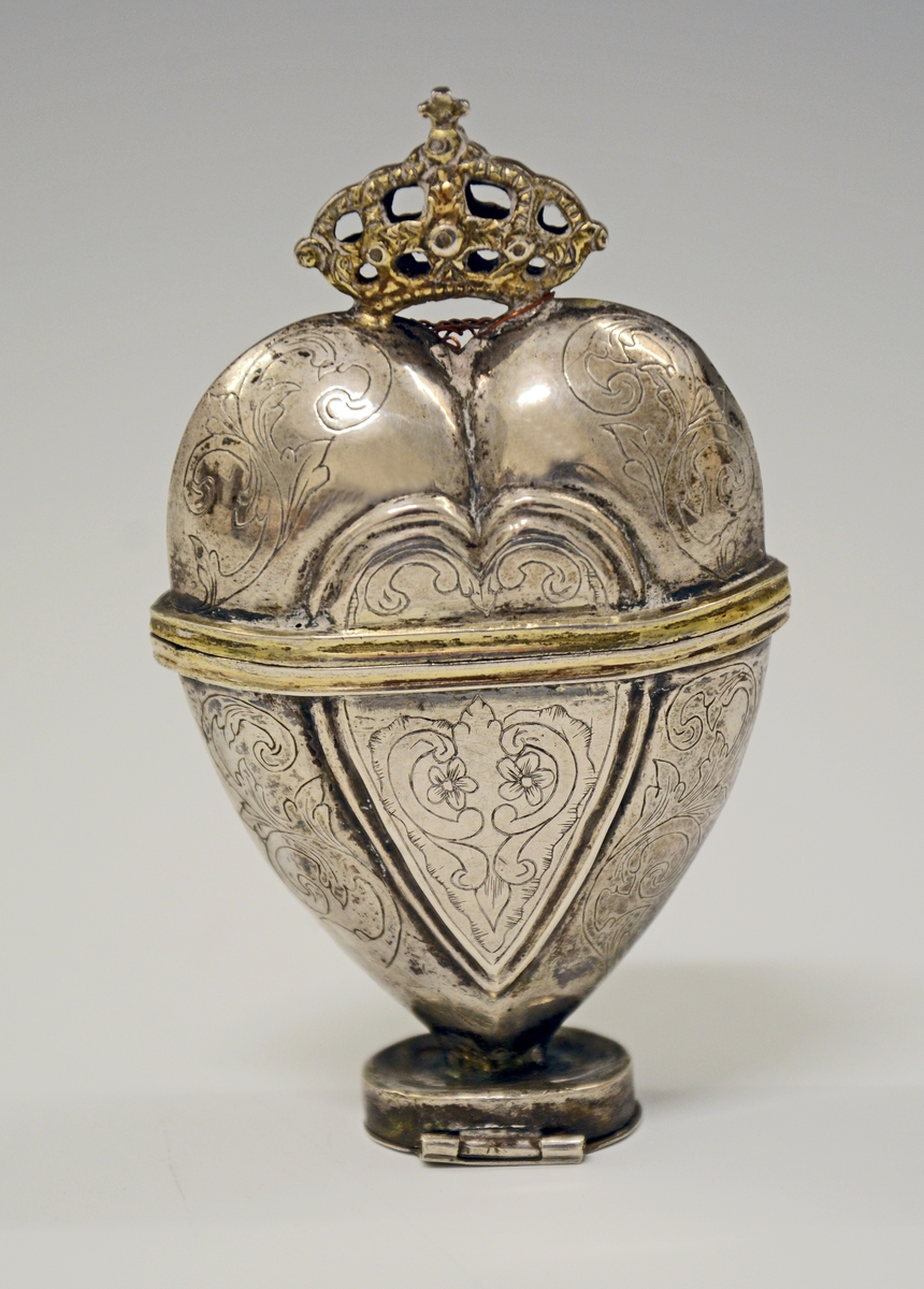 Fra protokollen:
Hjerteformet svampehus av sølv, bekronet, graverede ornamenter. Aapnes savel midtpaa som under fotsaalen.