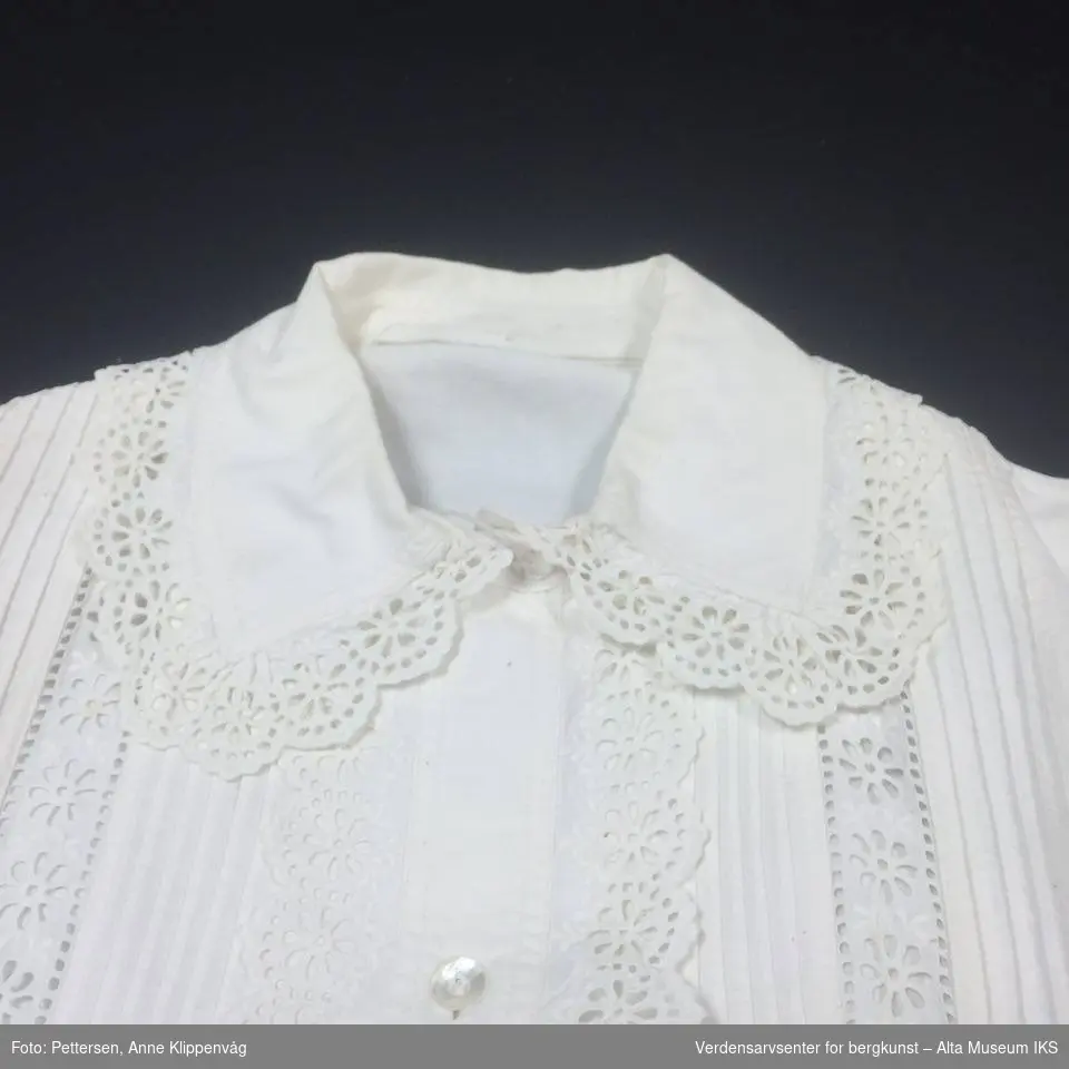 Langermet hvit skjorte med blonder rundt ermer, krage og ned langs forsiden.  Forknappet kvinneplagg med biser og blonder