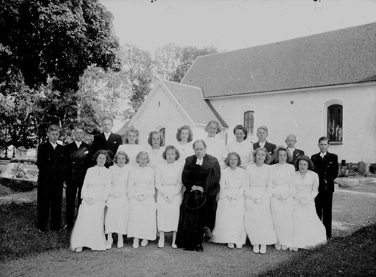 Konfirmander, 13 flickor, 6 pojkar och kyrkoherde Bror Strömberg.
Almby kyrka i bakgrunden.