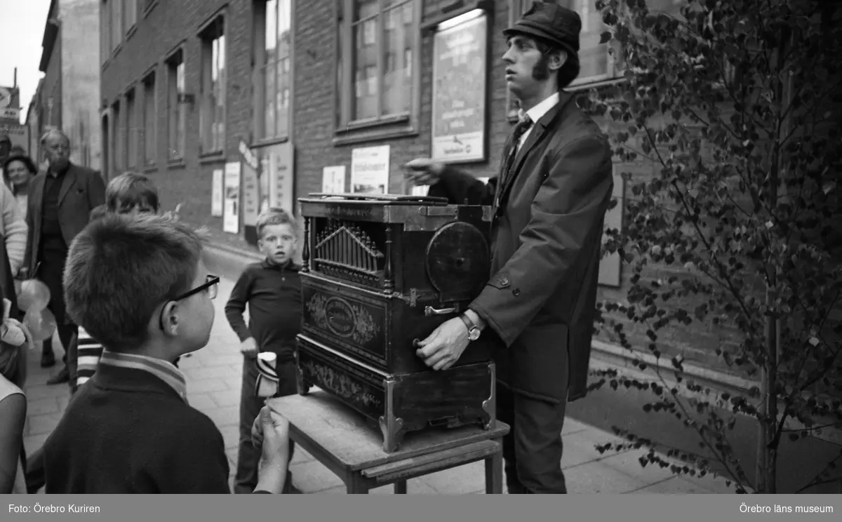 Marknadsafton 13 juni 1969

En man i jacka och hatt spelar på ett positiv på trottoaren. Några barn och vuxna ser på.