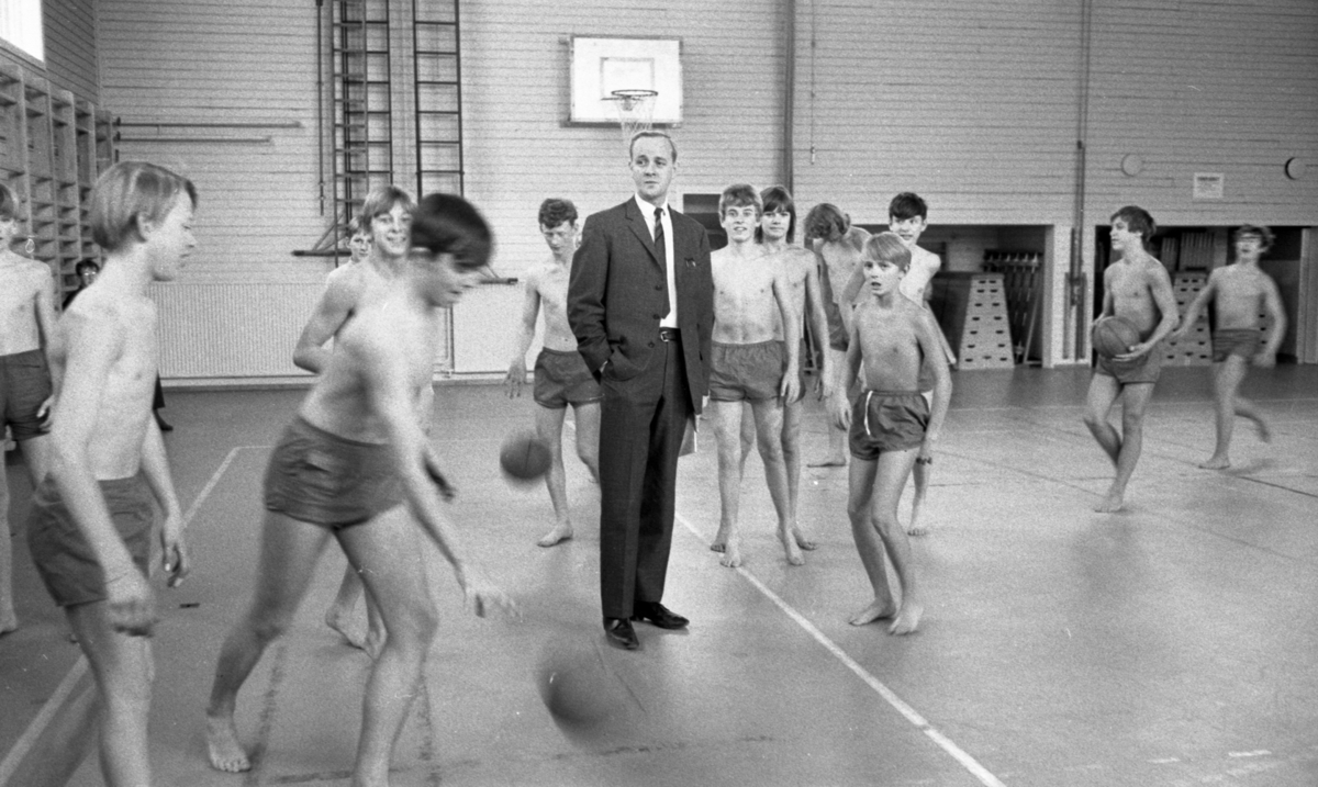 Mingporslin, Maranata, Strejken 25 oktober 1966

I en gymnastiksal på en skola spelar ett antal pojkar i fjortonårsåldern bollspel klädda i ljusa shorts. En vuxen man klädd i ljusgrå kostym, vit skjorta och ljusrandigt grå slips står mitt ibland dem. Plintar, basketkorg och ribbstolar syns bl.a. i bakgrunden.
