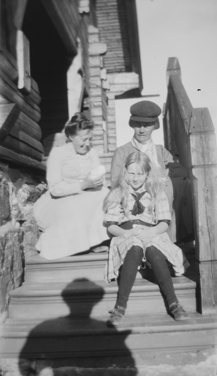 To damer og en jente sitter på en trapp av tre. Sola skinner.