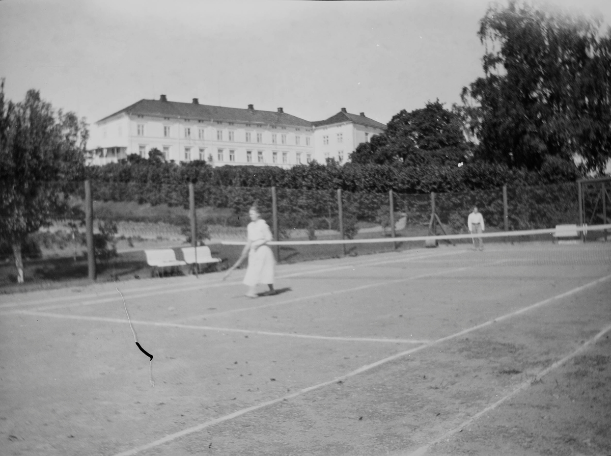 Tennisbane på Linderud gård om sommeren. En mann og en kvinne spiller tennis. I bakgrunnen; hage og hovedbygning.