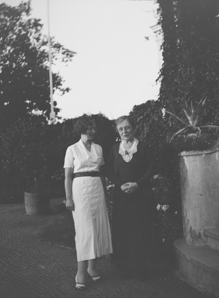 To kvinner står på hagestien ved siden av trappen opp til verandaen fra "Victorianske hagen"på Linderud Gård. Verandaen og muren som avgrenser hagen fra gårdsplassen er dekket av klatreplanter. Elsa Vetlesen til venstre og hennes mor, Marie Ihlen Vetlesen Gleditsch, til høyre.