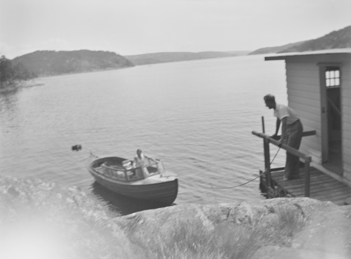 En mann lener seg mot et rekkverk på en brygge utenfor et "badehus" helt nede i sjøkanten. En snekke ligger fortøyd nedenfor. En kvinne står foran i båten vendt mot mannen på bryggen.
