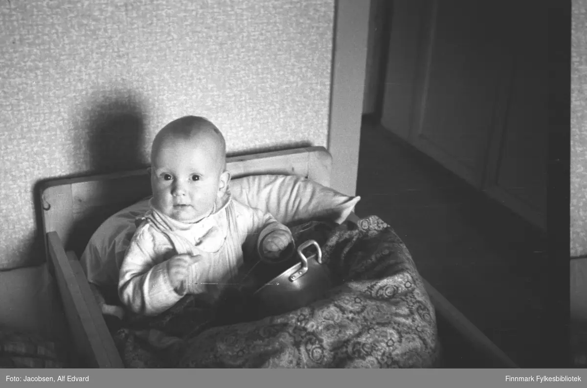 Arne Jacobsen sitter i sengen sin i Tromsø hvor familien bodde fram til sommeren 1947. Han har en hvit/lys skjorte på seg og noe av tøy rundt halsen, kanskje en smikke. Puten bak ryggen hans er stor og hvit. Sengeteppet/dyna er noe mørker og har heldekkende mønster. Foran han, på dyna, ligger en blank kasserolle med blankt håndtak. Han holder noe i sin venstre hånd, muligens en skje som han bruker som trommestikke. Barnesengen hans er trefarget og har litt høyre gavl enn sidene. Tapetet bak er ganske lyst og har et slags spraglet mønster. En døråpning med ganske lys dørlist og en lys speildør ses til høyre på bildet.