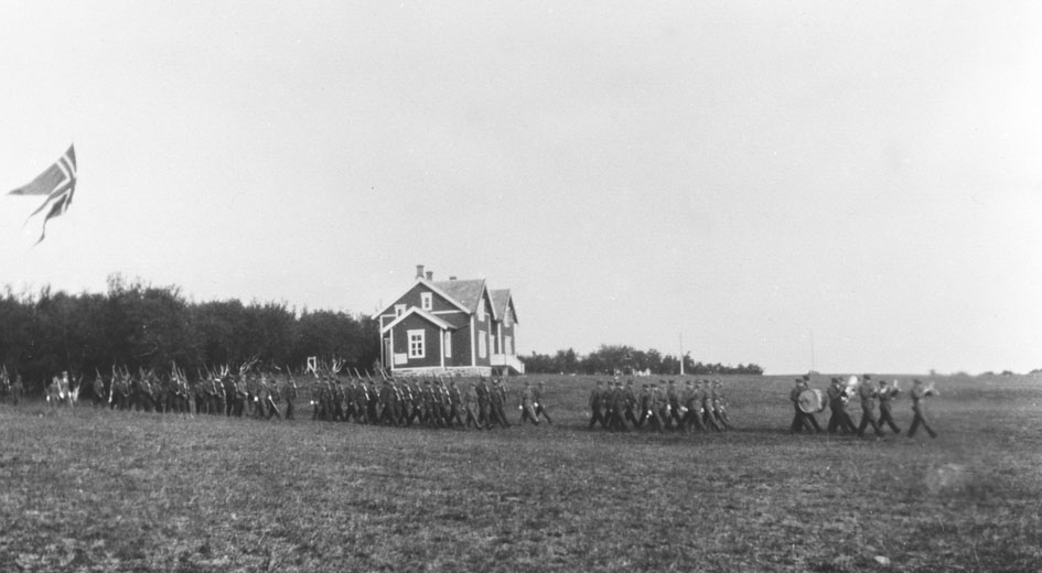 Vi antar at bildene er tatt i forbindelse med gjenopprettelse av Varanger batlajon 01.07.1934. Bildet viser marsjerende soldater ledet av militærkorps på eksersersplassen på Nybergmoen 1934. Soldaterhjemmet i bakgrunnen.