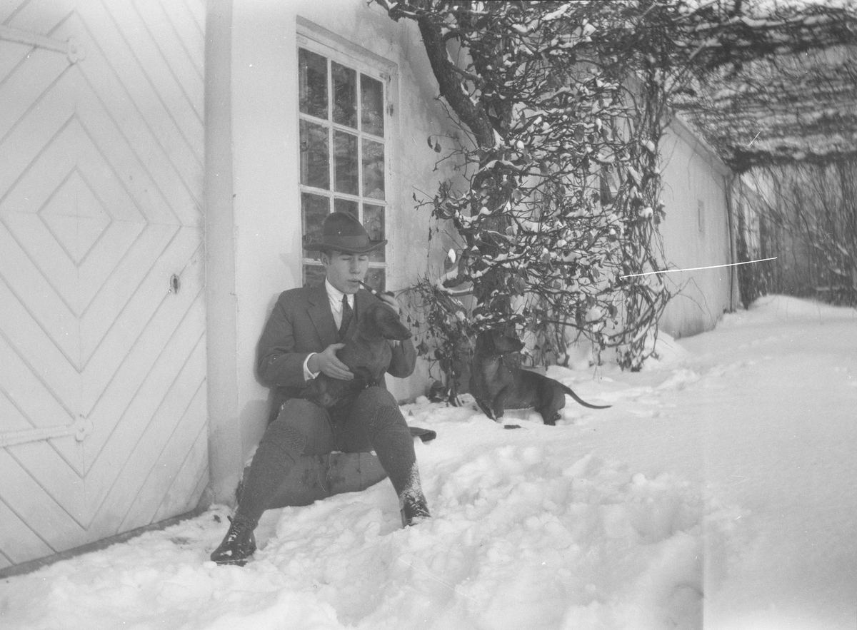 Iacob Ihlen Mathiesen sitter, på en stein eller lignende, utenfor en dør i grunnmuren til hovedhuset på Linderud Gård, med ham to dachser.