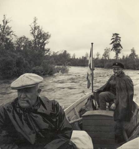 Två rökande män i en båt på strömt vatten. Buskvegetation längs stränderna. Mulet väder. Sommar.