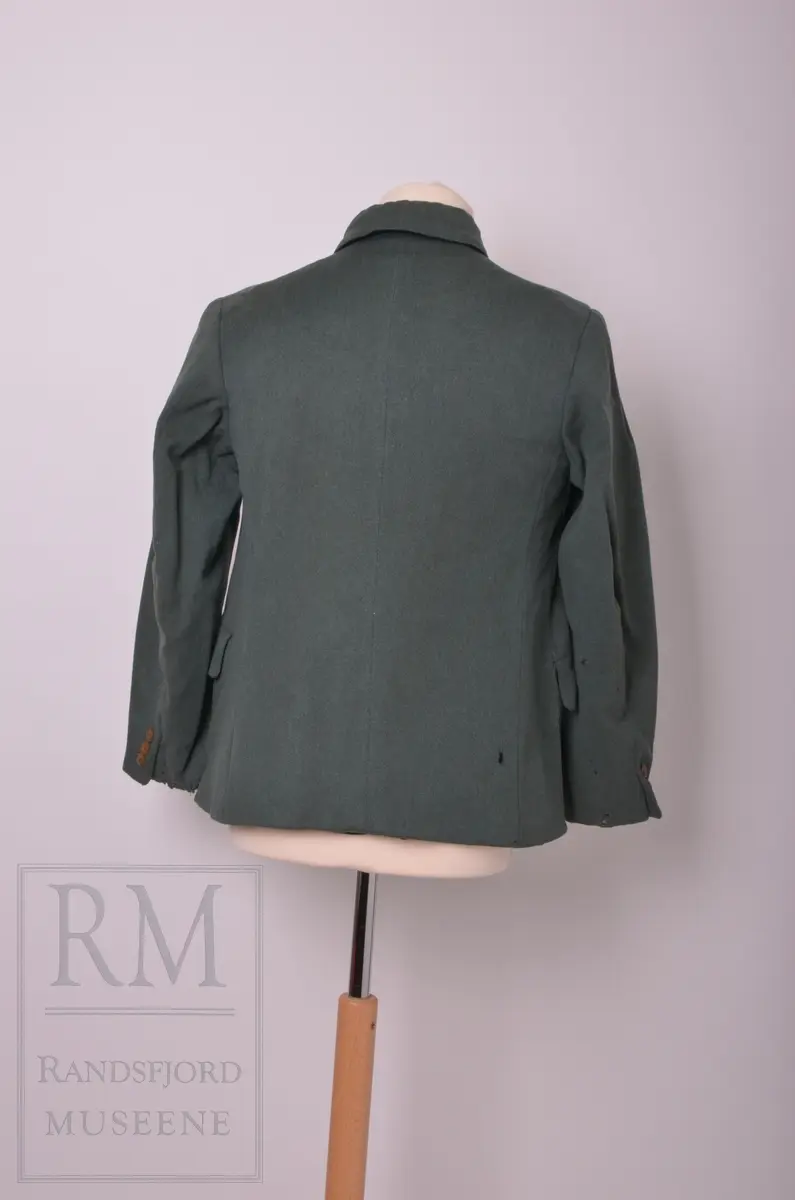 Mørk grønn jakke med svak A-fasong. Skjult knappestolpe og høy krave med korte snipper. Ikke originale knapper.