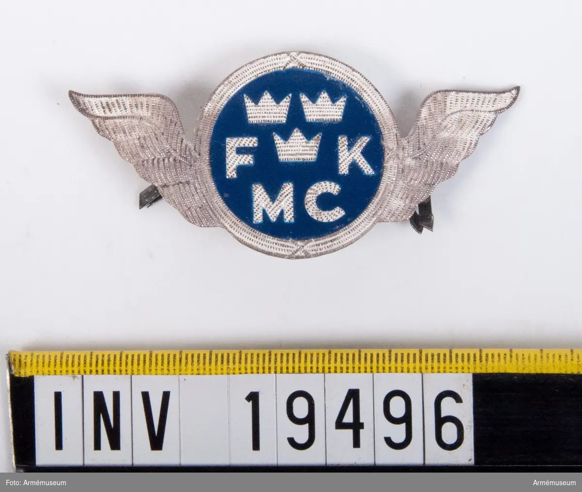 Tillverkat i aluminium. I mitten en blåmålad rundel varifrån två vingar utgår. I mitten trekronorsmärket och bokstäverna FK och MC finns på sidan om trekronorsmärket och nedanför. På baksidan finns en fästanordning.
