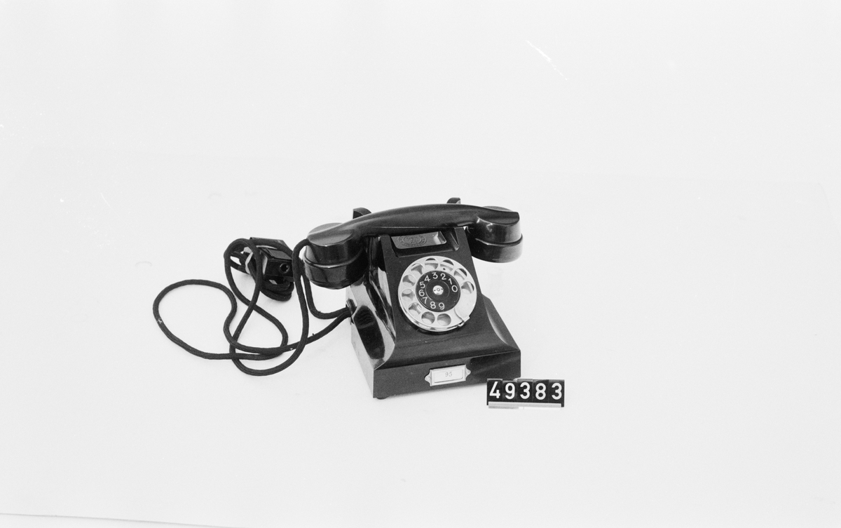 Telefonapparat BC 330, ändstationsapparat för AT-system. Bordapparat modell m33 av svart bakelit med fingerskiva av förnicklad mässing och textilklätt apparatsnöre anslutet till väggplint med lock av svartlackerad plåt.