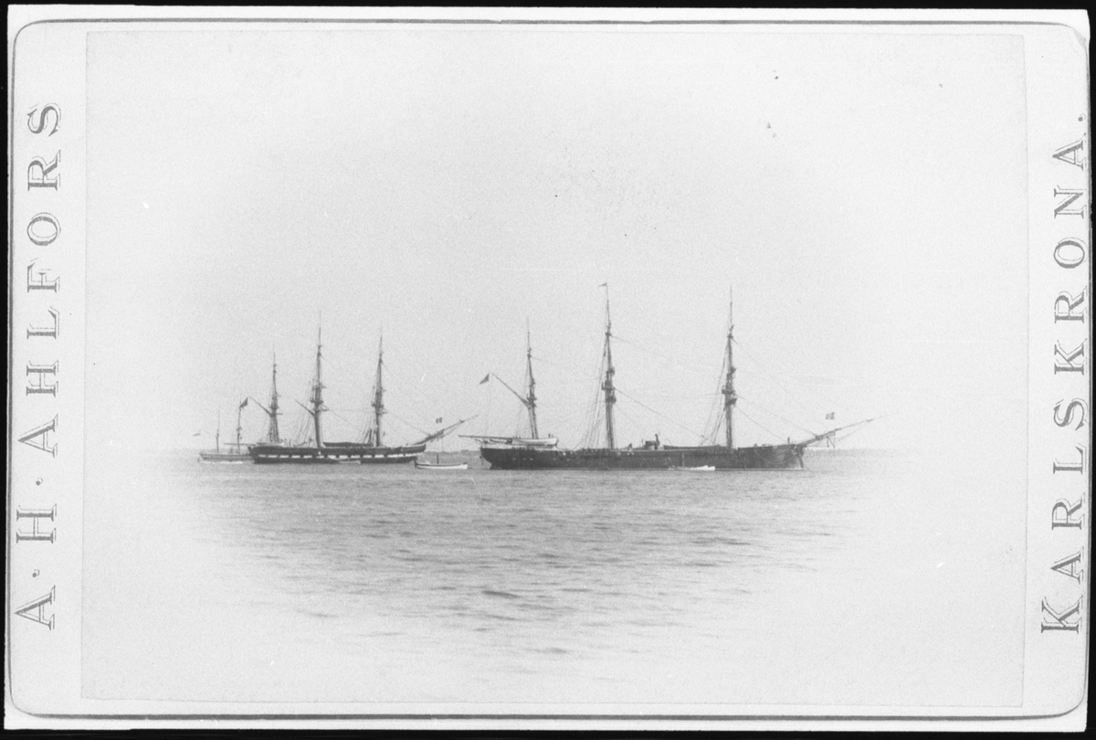 Reprofotografi efter ett vykort som visar fregatten Norrköping och korvetten Saga som ankrar i Karlskrona. Vänster om Norrköping ligger en tredje segelfartyg. Före skeppen syns flera mindre roddbåtar.
