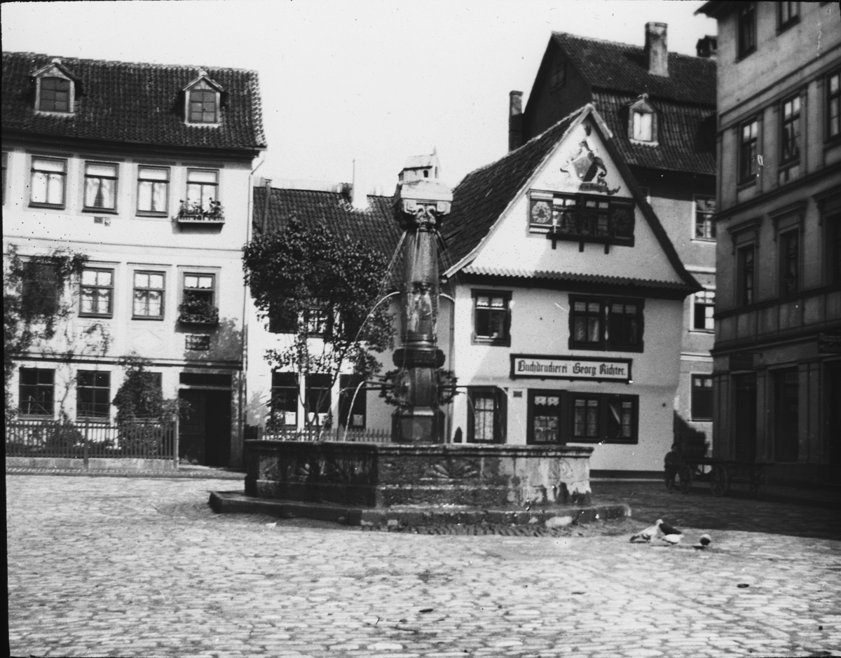 Skioptikonbild med motiv av fontänen Kapellenbrunnen på torget Platz an der Kapelle i Meiningen.
Bilden har förvarats i kartong märkt: Vårresan 1910. Weimar 3. Meiningen 3. IV.