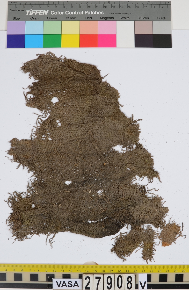 Textil.
Ca 263 textilfragment uppdelade på fyndnummer 27908:I-XII. De romerska siffrorna står för lagerföljden, d.v.s. den ordning i vilken textilen hittades, i svepask 13701.
Fnr 27908:I består av ca 15 fragment av ull vävda i tuskaft samt 2/2-kypert (fyrskaft). Beige tuskaftväv av ett ursprungligen grått garn med 11 tr z-spunnet garn per cm i ena riktningen och 14 tr s-spunnet garn per cm i andra riktningen. Fragmenten har flera originalkanter med sömmar med bevarad tråd kvar och ser ut att ha kunnat vara manschetter eller kragbitar till en skjorta eller jacka. Ett gråbeige kyperttyg har varit fastsytt på tuskafttyget. Kyperttyget är vävt av ett garn av vegetabiliska fibrer med 10 tr/cm i ena riktningen och ett ullgarn med 6 tr/cm i den andra riktningen. Ullgarnet är omväxlande grått och beige i färgen i ränder. Ovanpå det vävda lagret finns ett tunt lager vegetabiliska beige trådar.
Fnr 27908:II består av ca 62 fragment. Ett fragment är vävt i tuskaft av 10 tr z-spunnet x 8 tr s-spunnet grått garn per cm. Det har varit valkat. Övriga fragment är samma typ av 2/2-kypert som i lager I. Lager II innehåller även några mindre fragment av samma vegetabiliska väv som finns i lager XI, med ett gulgrönt korrosionsmärke på ett fragment, eventuellt efter en knapp.
Fnr 27908:III består av ca 35 fragment i 2/2-kypert (fyrskaft) av z-spunnet grått garn. Garnet är troligen av ull men väldigt sprött. En del av fragmenten har bevarade fållar.
Fnr 27908:IV består av 2 fragment i samma 2/2-kypert som i lager III. Det mindre fragmentet har en bevarad fåll.   
Fnr 27908:V består av 3 fragment i samma 2/2-kypert som i lager III. 
Fnr 27908:VI består av ca 12 fragment i samma 2/2-kypert som i lager III.  Det större fragmentet har en bevarad fåll.
Fnr 27908:VII består av 3 fragment i samma 2/2-kypert som i lager III. 
Kypertväven i lager III-VII liknar varandra.
Fnr 27908:VIII består av ca 28 fragment i kypert. Ull eller lin? Kypertväv, eventuellt samma som i lager III-VII ovan men i så fall mycket mer ihoppressad. Kan också vara samma vegetabiliska väv som i lager XI.
Fnr 27908:IX består av ca 22 fragment. 18 av fragmenten är av vitbeige ull och nålbundna (av S-spunnet tvåtrådigt garn), men 4 fragment är beigebruna och vävda.
Fnr 27908:X består av ca 54 fragment varav ca 30 fragment är nålbundna av vitbeige ull som ovan + 18 gråbeige kypertfragment + 5 beigebruna kypertfragment + 1 beigebrunt fragment i tuskaft med hål efter söm längs ena kanten. Tuskaftsväven i lager X hör troligen till en av varianterna i lager I eller II.
Fnr 27908:XI består av totalt 24 textilfragment samt 3 fragment av en knapp. Innehåller ett större fragment med stomme av brunt ullgarn, stickat eller vävt? Väv av vegetabiliska fibrer av z-spunnet garn med 9x7 tr/cm ovanpå det. 19 små tygfragment har lossnat från föremålet. Fortfarande fast på tyget sitter 13 knapphål, sydda med langettstygn av tvåtrådig beige silkestråd. Det finns också en lös rad bestående av 5 sammanhängande knapphål av samma sort som de andra. Innanför de lösa knapphålens tråd skymtar rester av tyg. Knappfragmenten består av två halvor av en metallknapp samt knappens metallögla fanns också med i fyndasken. Fyndnumret innehåller även ett band vävt i tuskaft av grått silke med 2-tr garn i varpen och 1-tr garn i inslaget, 18 dubblatr/cm samt två garndockor av 2-tr s-tvinnat grått ullgarn med tvinnat silkestråd runt.  
Fnr 27908:XII består av 4 slätstickade ullfragment, varav två är rundstickade av dels ljust och dels grått garn i randning, 1-tr s-tvinnat med 2 maskor/cm och 2 varv/cm. Lagret innehåller även 3 hårdare små fragment av oklart material.