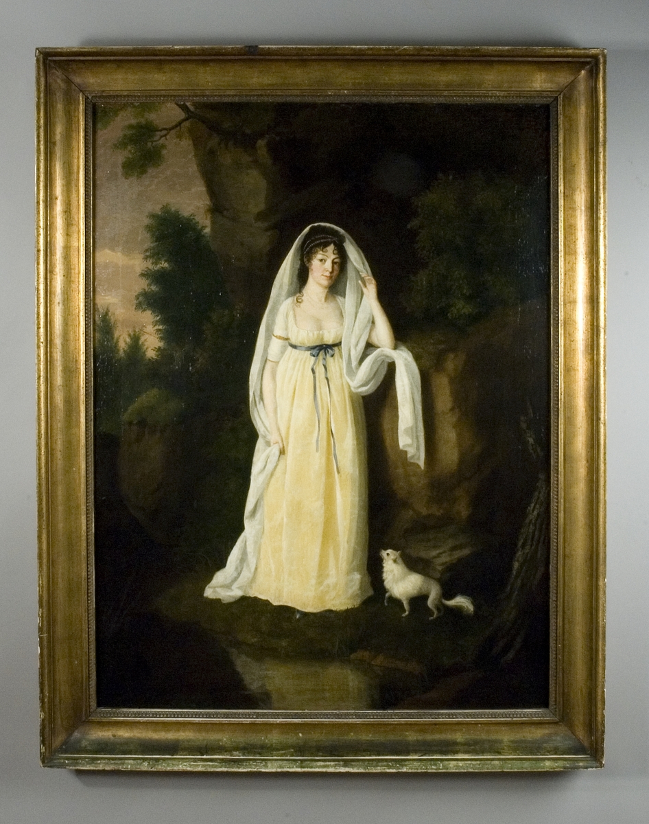 Porträtt av kvinna i helfigur, Fredrika Charlotta Stenbock, Lolotte, klädd i  gulvit empireklänning stående vid en klippa. Vid hennes fötter en liten vit hund. Skog i bakgrunden.